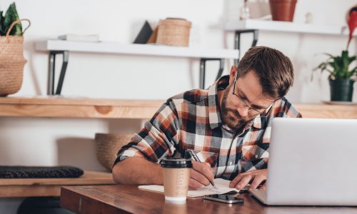 Focused young entrepreneur sitting at a table at home writing in a notebook next to a laptop